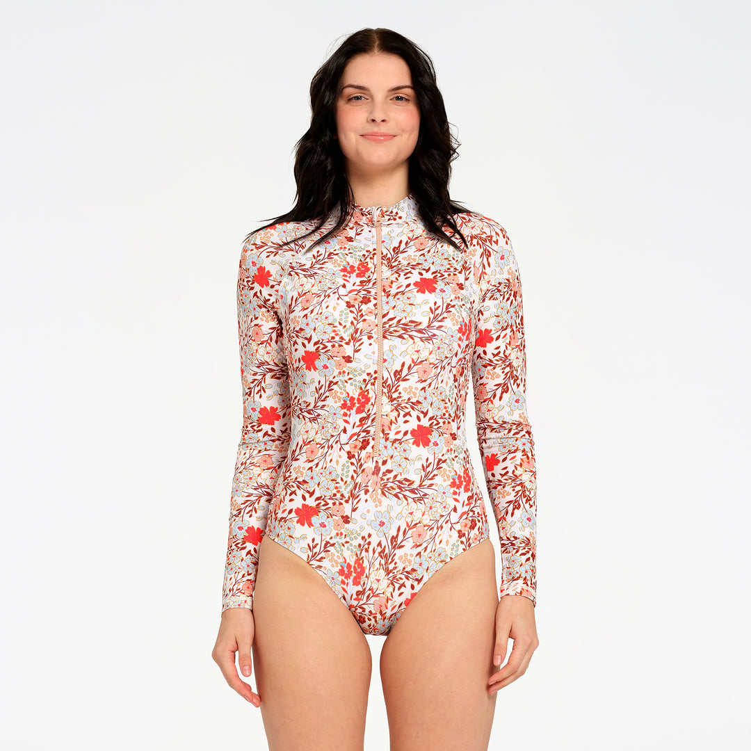A woman modeling a long-sleeve high-neck one-piece swimsuit with a red and orange floral pattern on a white background, set against a plain light gray studio backdrop.