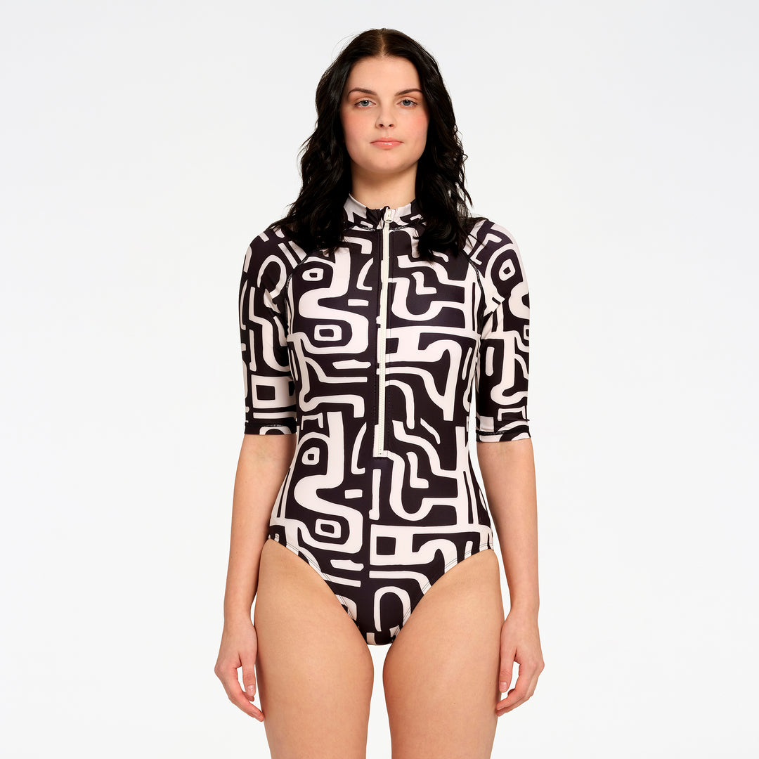 A woman modeling a short-sleeve one-piece swimsuit with a black and off-white abstract maze-like pattern, a front zipper, and a high collar, set against a plain white studio background.