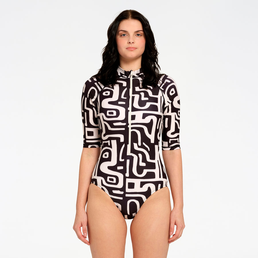 A woman modeling a short-sleeve one-piece swimsuit with a black and off-white abstract maze-like pattern, a front zipper, and a high collar, set against a plain white studio background.