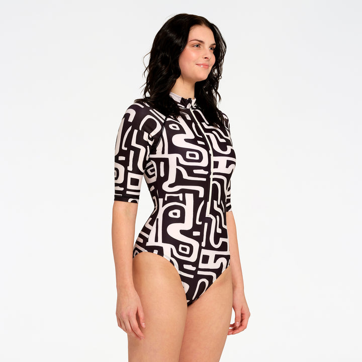 A woman modeling a short-sleeve one-piece swimsuit with a black and white abstract geometric pattern, photographed against a solid white studio background.
