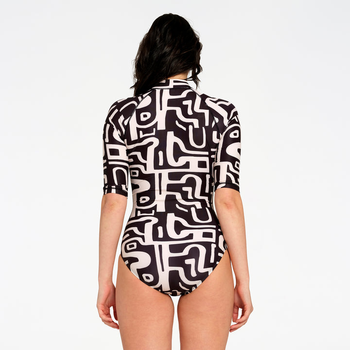 A woman viewed from the back wearing a short-sleeve one-piece swimsuit with a bold black and white abstract maze-like pattern, set against a plain white studio background.
