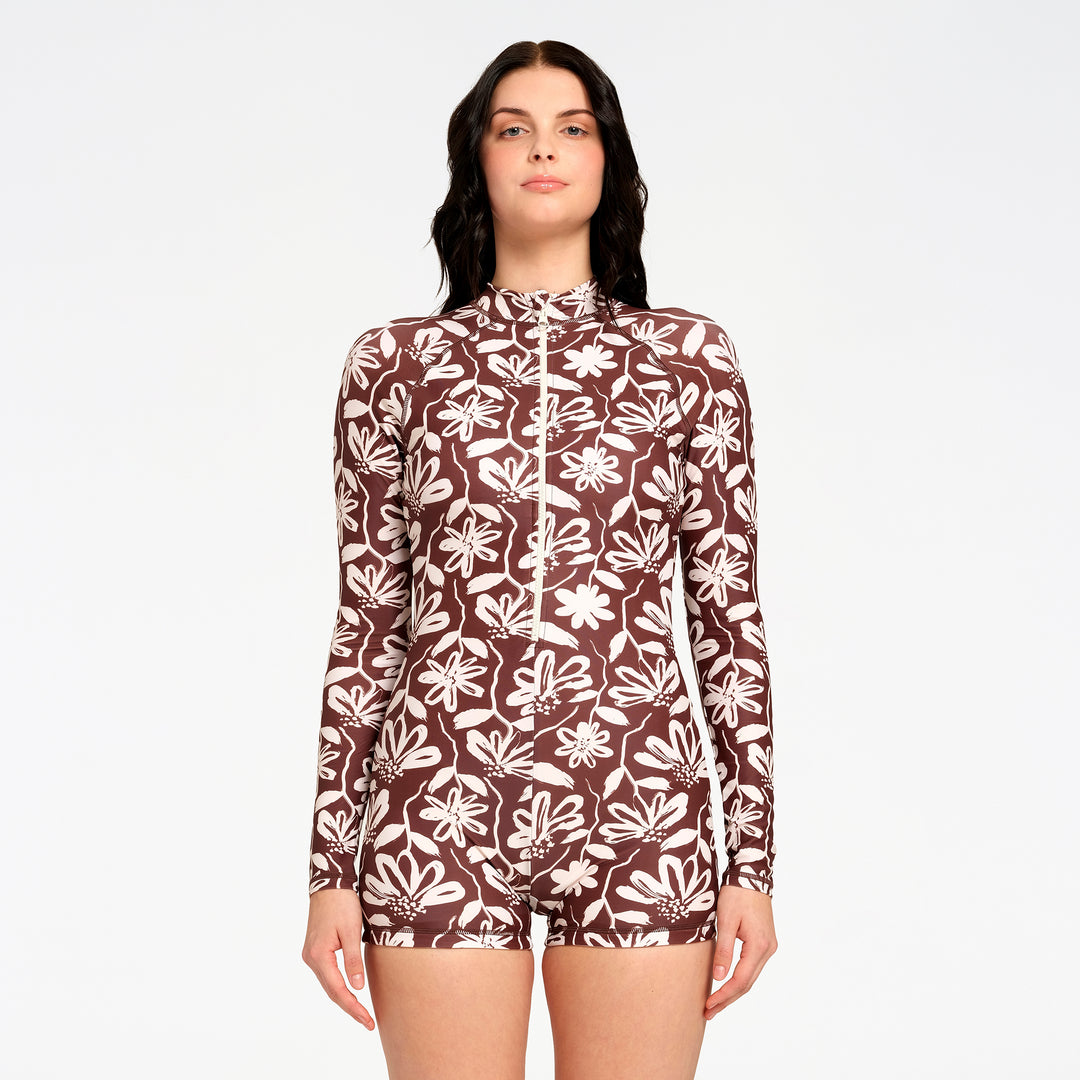 A woman modeling a long-sleeve, high-neck one-piece swimsuit with a brown and off-white floral pattern, set against a plain light gray background.