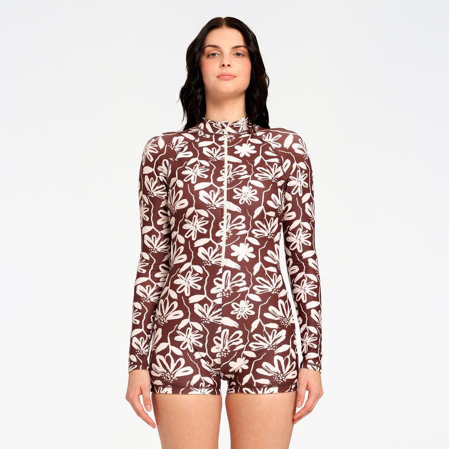 A woman modeling a long-sleeve, high-neck one-piece swimsuit with a brown and off-white floral pattern, set against a plain light gray background.