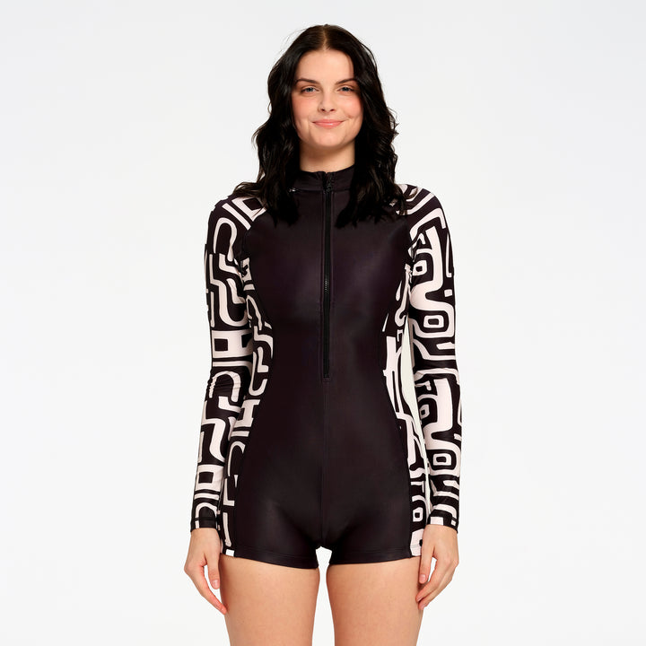 A woman modeling a black and white patterned long-sleeve one-piece swimsuit with a front zipper, set against a plain white studio background.