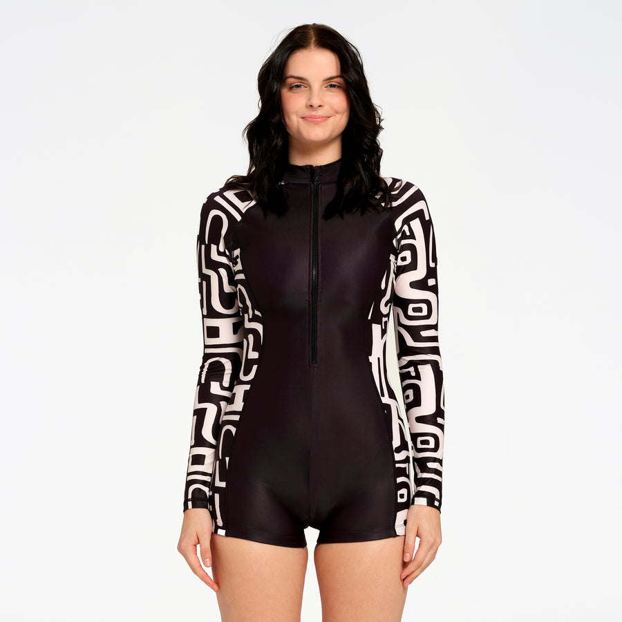 A woman modeling a black and white patterned long-sleeve one-piece swimsuit with a front zipper, set against a plain white studio background.