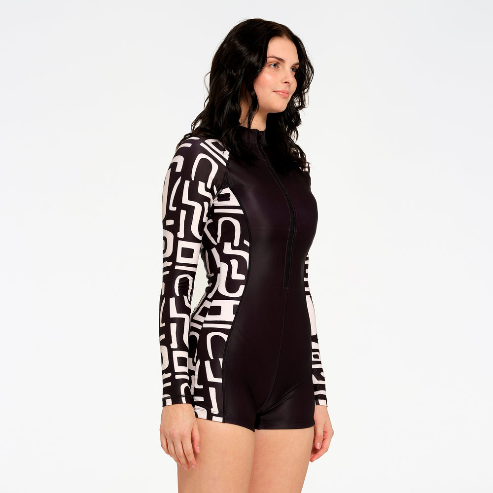 A woman modeling a one-piece long-sleeve swimsuit with a black and white geometric pattern on the sleeves and sides, and a solid black front torso, against a plain white studio background.