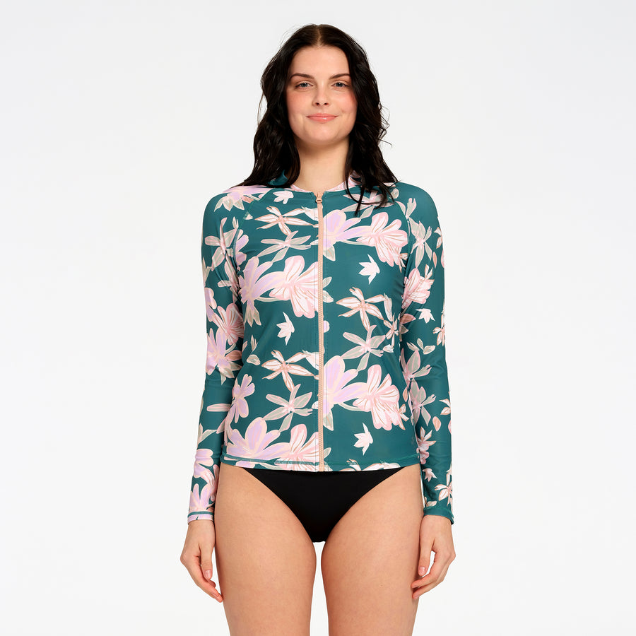 A woman modeling a long-sleeve swim rash guard with a teal green background and a pink and beige floral pattern against a plain white studio background.