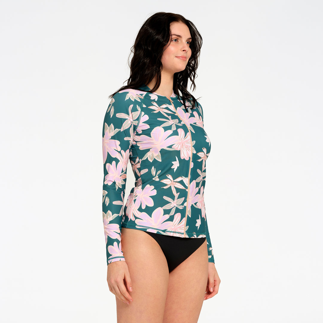 A woman modeling a long-sleeve swim rash guard featuring a teal green background with a pink and white floral pattern against a plain white studio background.