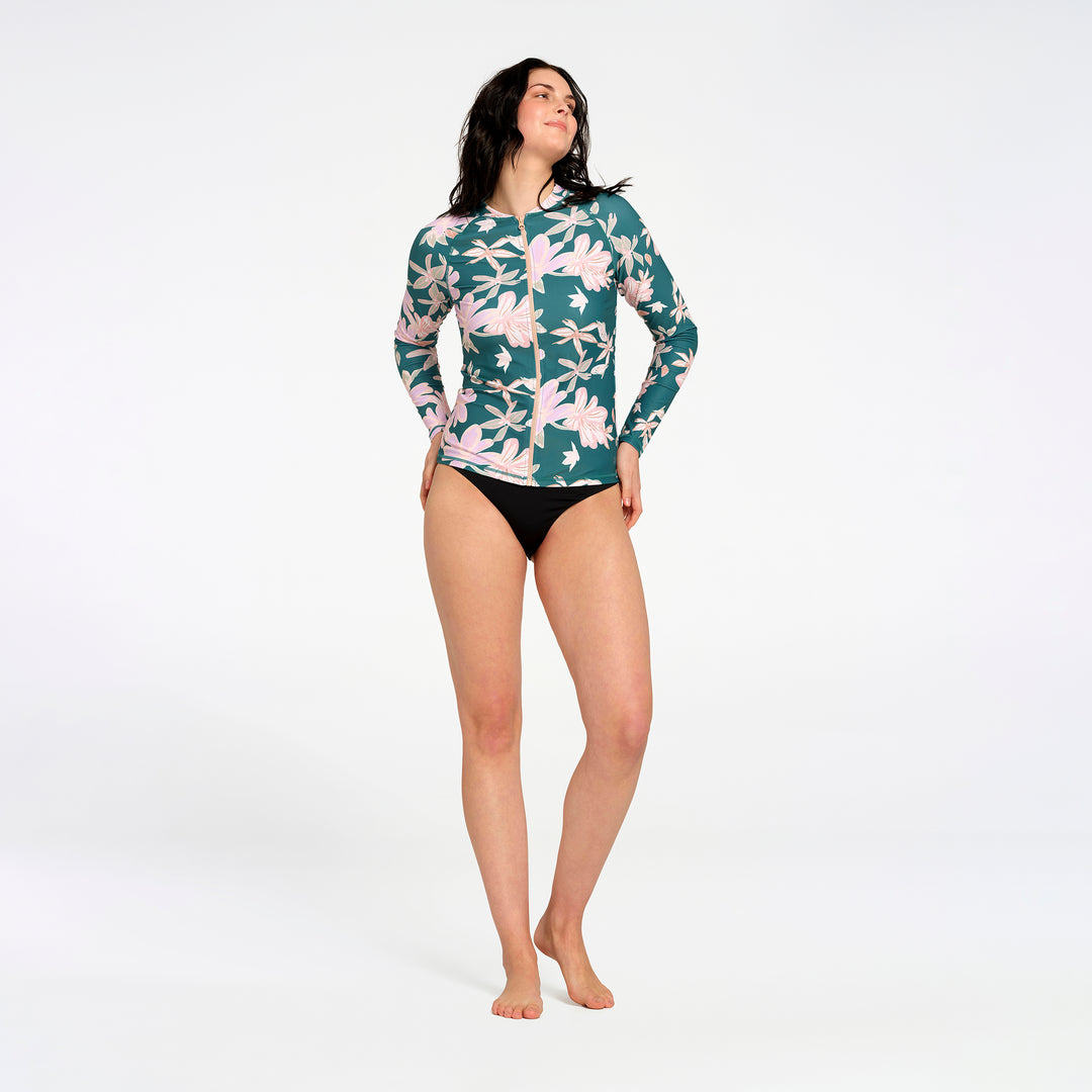 A woman modeling a long-sleeve swim rash guard with a green, pink, and white floral pattern, against a plain white studio background.