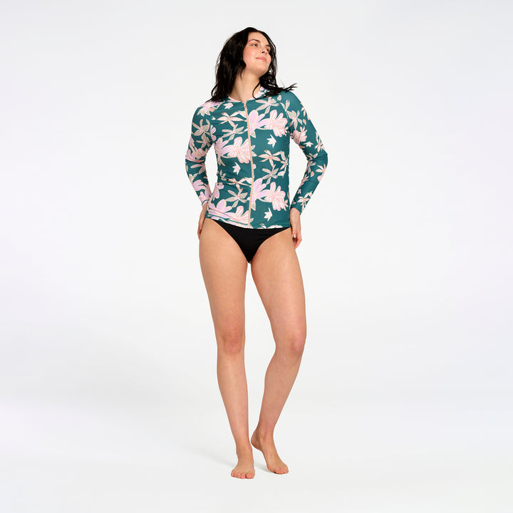 A woman modeling a long-sleeve swim rash guard with a green, pink, and white floral pattern, against a plain white studio background.