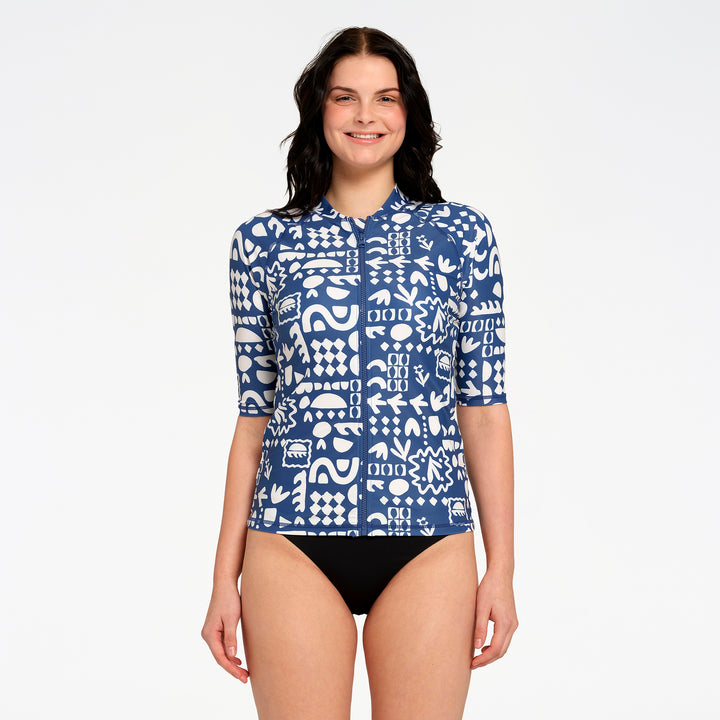 A woman modeling a blue and white patterned swim top with three-quarter sleeves, paired with black swim bottoms, against a plain white studio background.