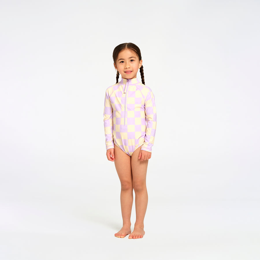 A young girl with braided hair stands barefoot against a white studio background, wearing a long-sleeve one-piece swimsuit with a light purple and pale yellow geometric pattern and a front zipper.