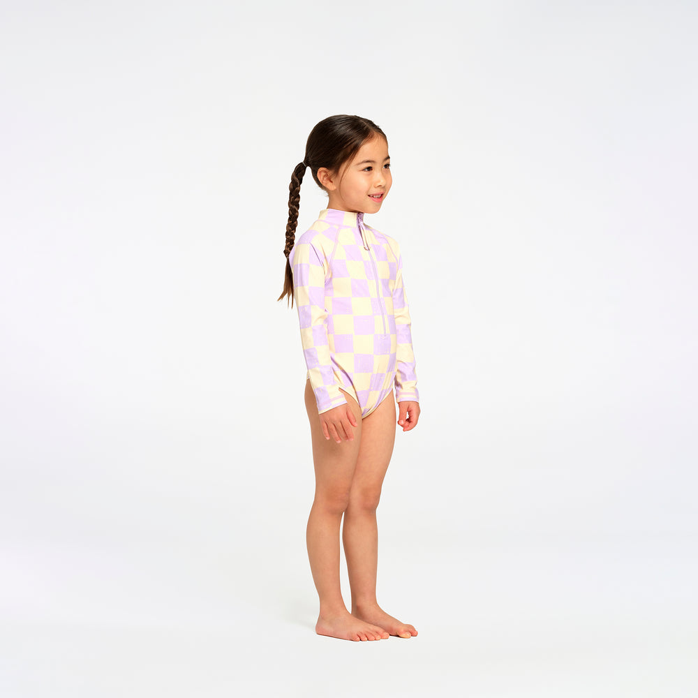 A young girl with a braided ponytail stands in profile wearing a long-sleeve one-piece swimsuit with a pastel lavender and yellow checkerboard pattern, set against a seamless white studio background.