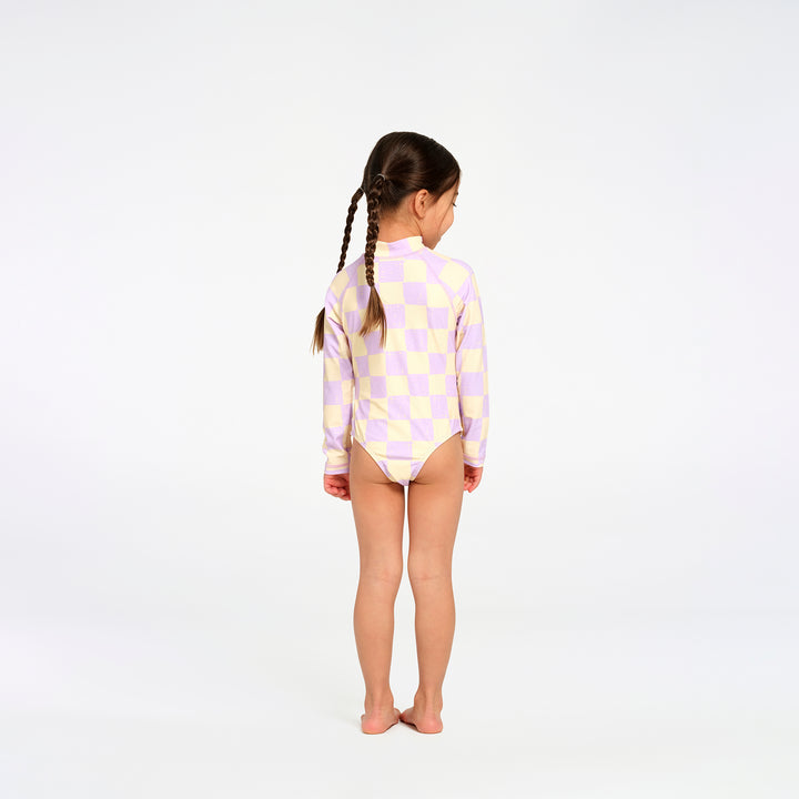 A young girl with braided hair stands with her back to the camera wearing a long-sleeve one-piece swimsuit with a pastel yellow and lavender checkerboard pattern, set against a plain white studio background.