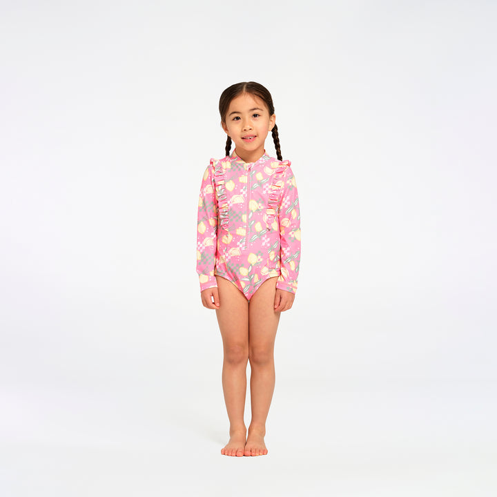 A young girl with braided hair stands barefoot against a seamless white studio background, wearing a pink long-sleeve swim suit with a floral and lemon print.