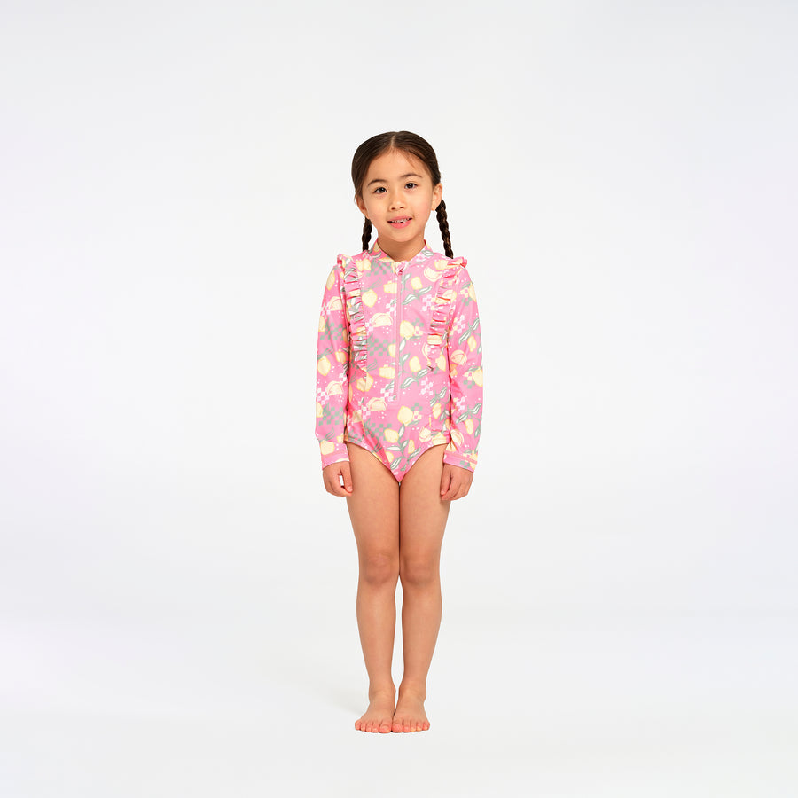 A young girl with braided hair stands barefoot against a seamless white studio background, wearing a pink long-sleeve swim suit with a floral and lemon print.