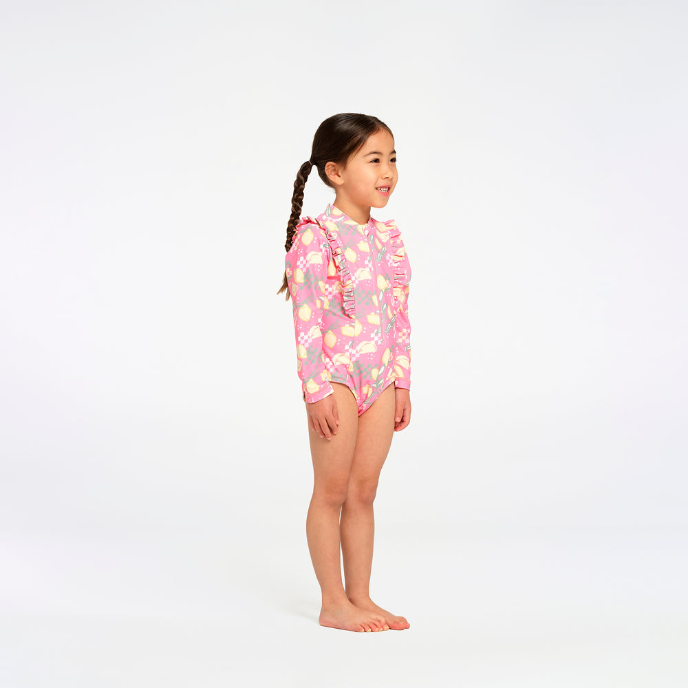 A young girl with braided hair stands in profile wearing a pink long-sleeve swim suit with a colorful floral pattern, set against a plain white studio background.