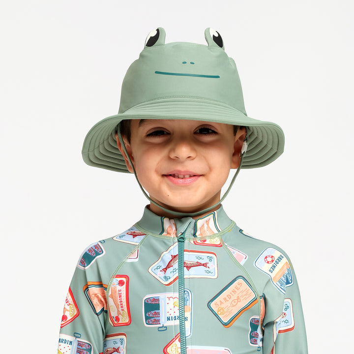A young child wearing a sage green frog-themed sun hat with a chin strap and a matching sage green long-sleeve swimsuit with a colorful sardine print