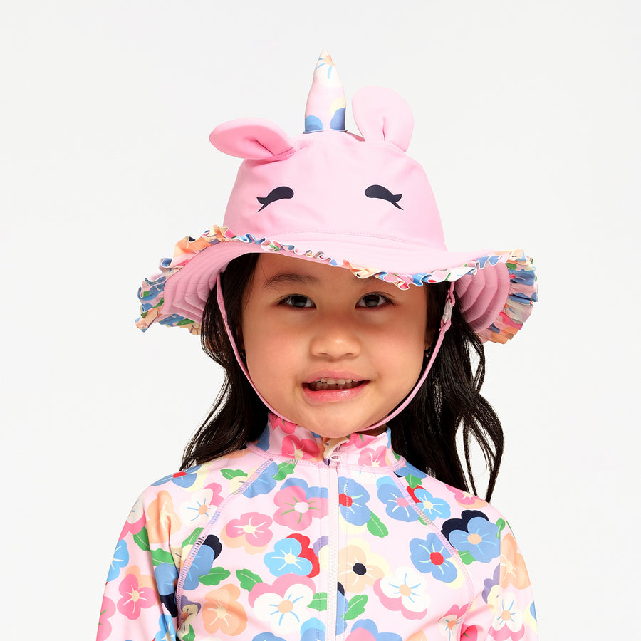A young child wearing a pink unicorn sun hat with a floral-patterned swimsuit, photographed against a clean white background.