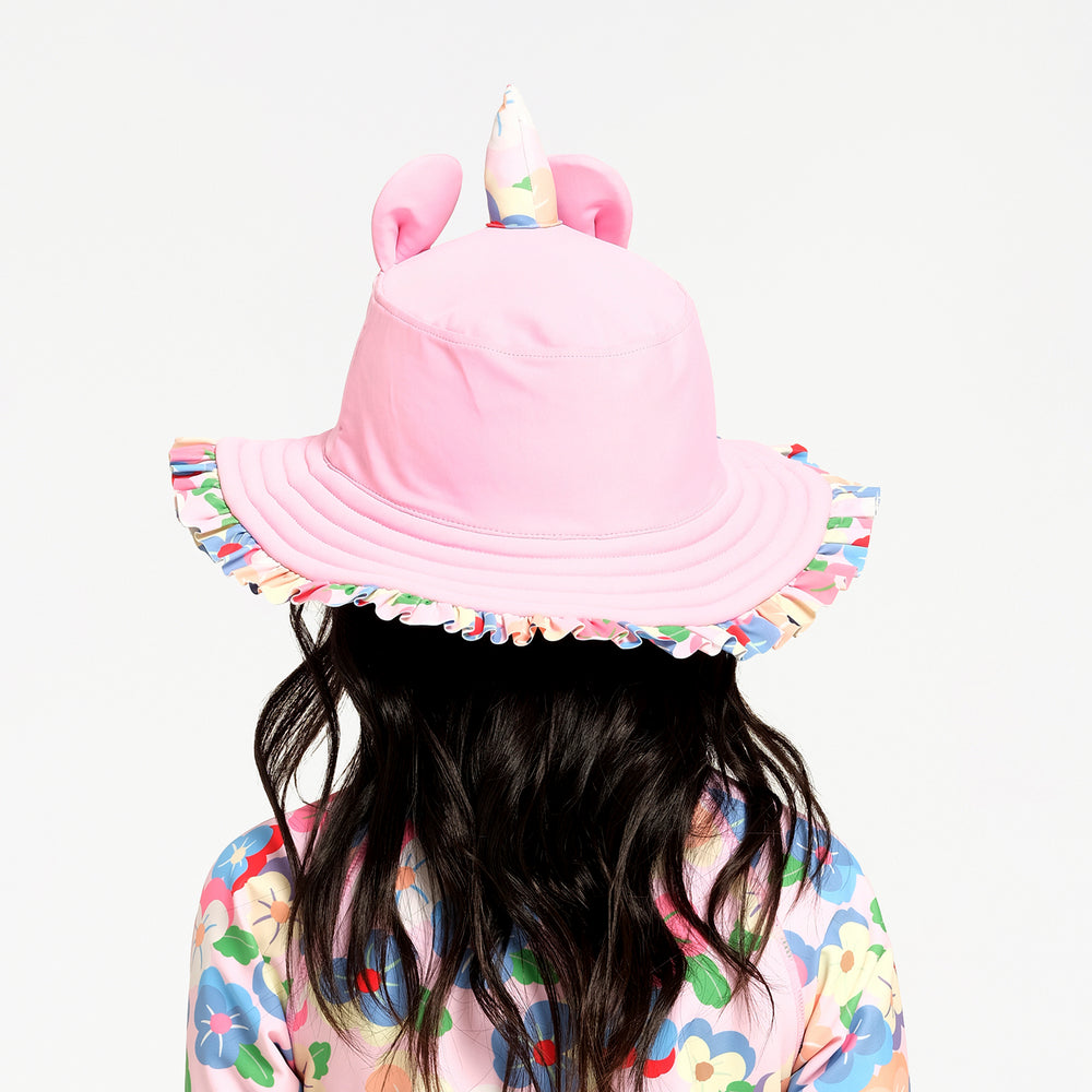 A child viewed from behind wearing a pink bucket hat with ears and a unicorn horn, featuring a multicolor floral ruffled brim, against a plain white background.
