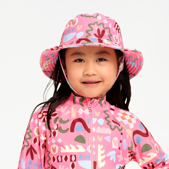 A young girl wearing a pink patterned sun hat with a chin strap and a matching pink patterned long-sleeve zip-up shirt, photographed against a clean white background.