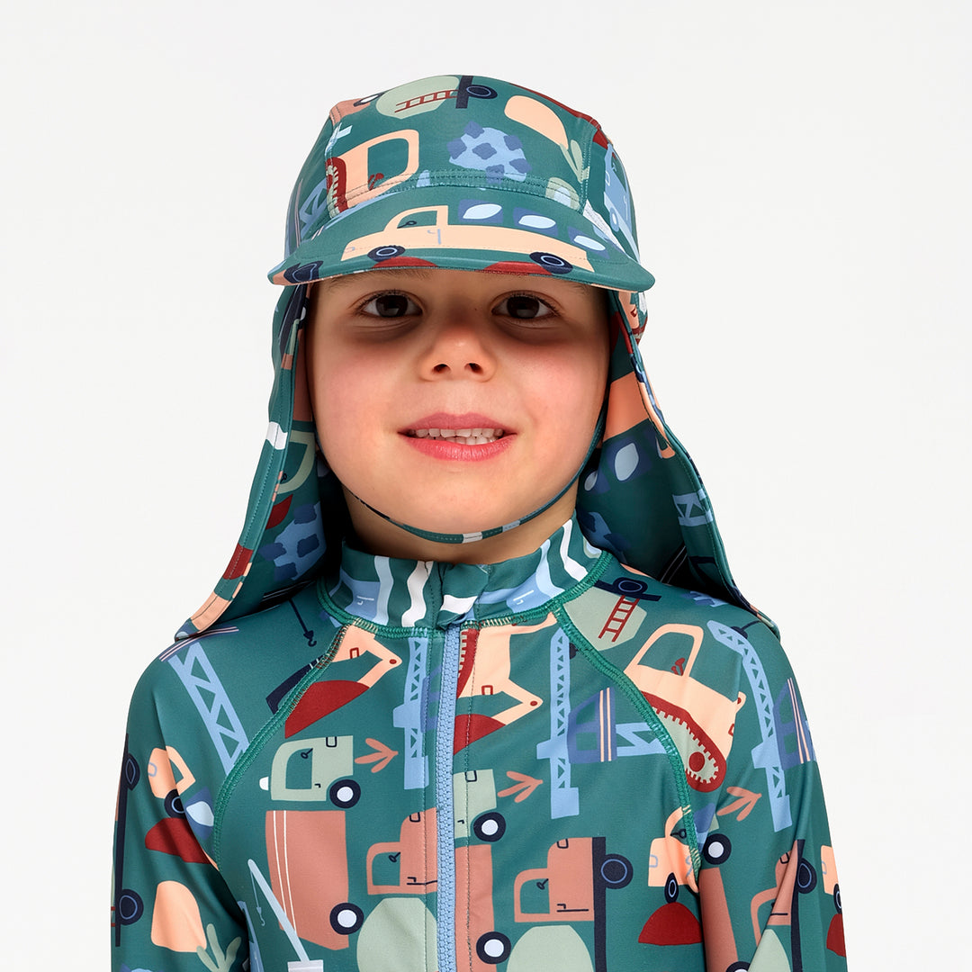 A child wearing a matching full-zip swimsuit and sun hat with a colorful construction vehicle print on a teal background, set against a plain light gray studio background.