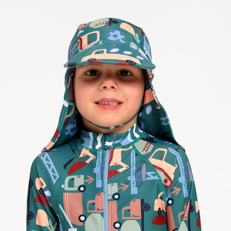 A child wearing a matching full-zip swimsuit and sun hat with a colorful construction vehicle print on a teal background, set against a plain light gray studio background.