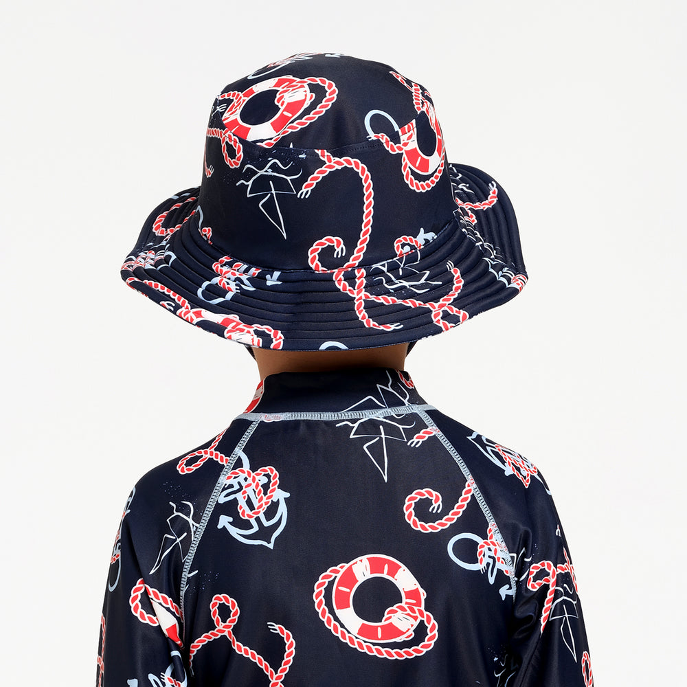 A child shown from the back wearing a matching navy blue sun hat and long-sleeve rash guard with a red and white rope, life preserver, and white anchor pattern, against a plain white background.