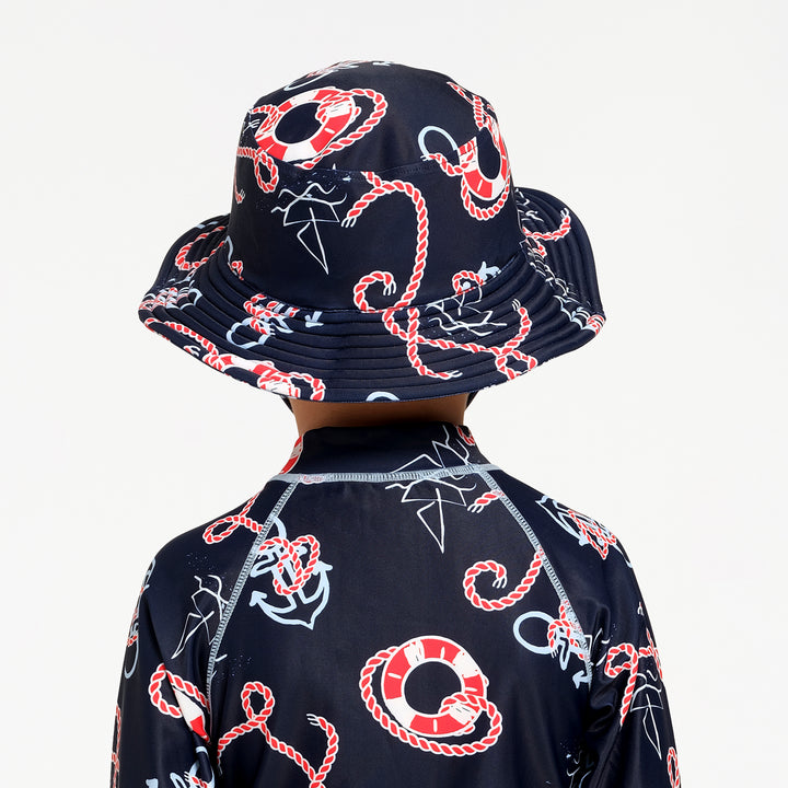 A child shown from the back wearing a matching navy blue sun hat and long-sleeve rash guard with a red and white rope, life preserver, and white anchor pattern, against a plain white background.