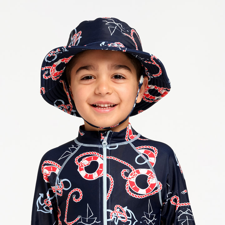 A young boy smiling while wearing a navy blue nautical-themed children's sun hat and matching zip-up swimsuit with red and white nautical prints, set against a plain light gray background.