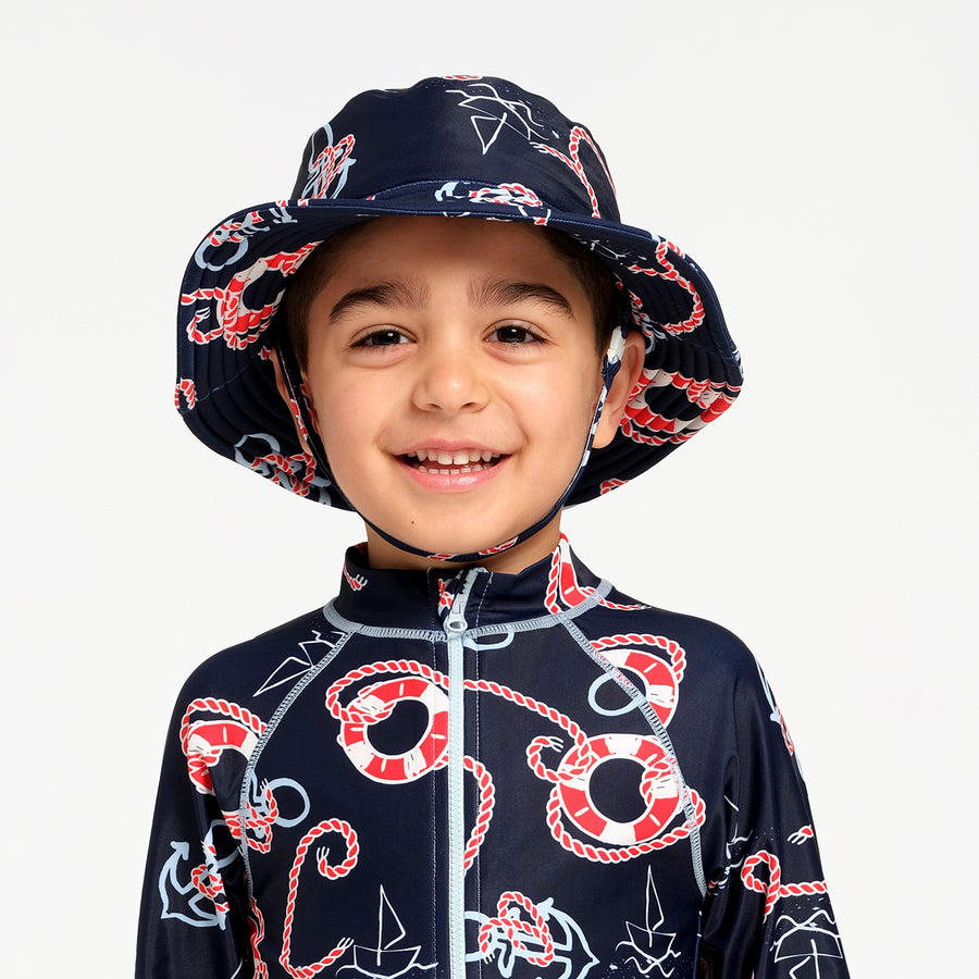 A young boy smiling while wearing a navy blue nautical-themed children's sun hat and matching zip-up swimsuit with red and white rope, life preservers, and sailboat prints, set against a plain light gray background.
