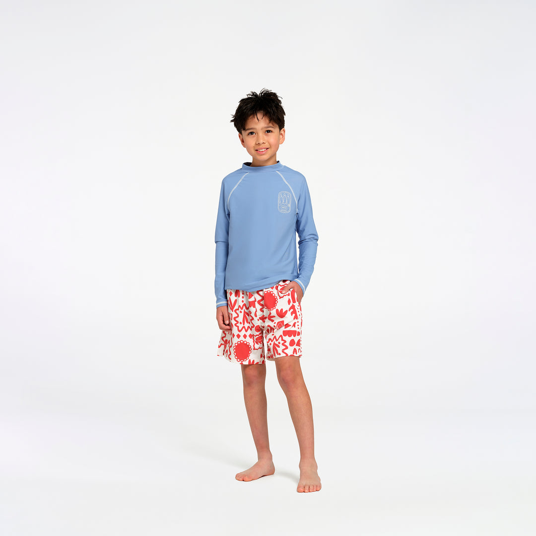 A young boy stands barefoot against a white studio background, wearing a light blue long-sleeve rash guard with a subtle chest logo and red-and-white floral print swim trunks.