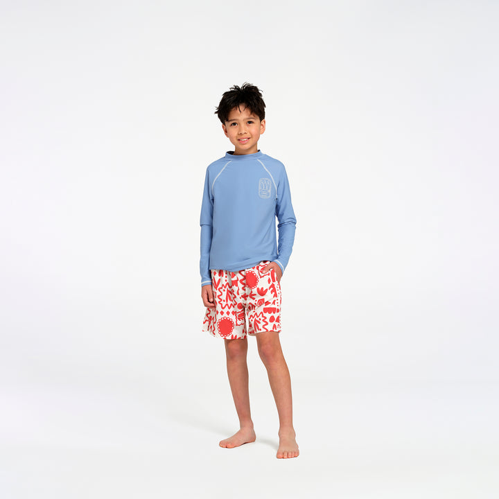 A young boy stands barefoot against a white studio background, wearing a light blue long-sleeve rash guard with a subtle chest logo and red-and-white floral print swim trunks.