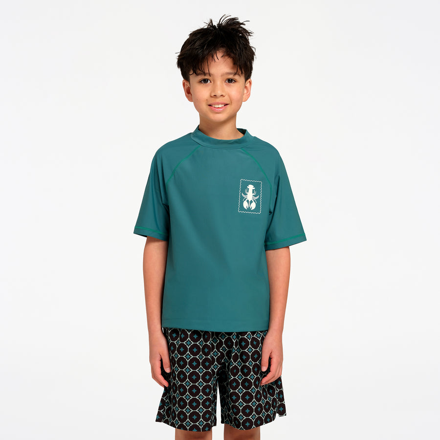 A young boy wearing a teal short-sleeve rash guard with a white chest logo and patterned swim shorts, standing against a plain white studio background.