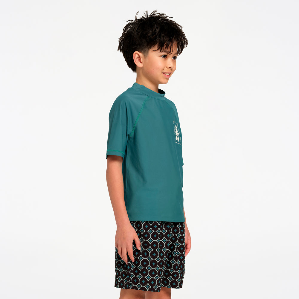 A young boy wearing a teal short-sleeve rash guard and patterned swim shorts, standing in profile against a solid white background.