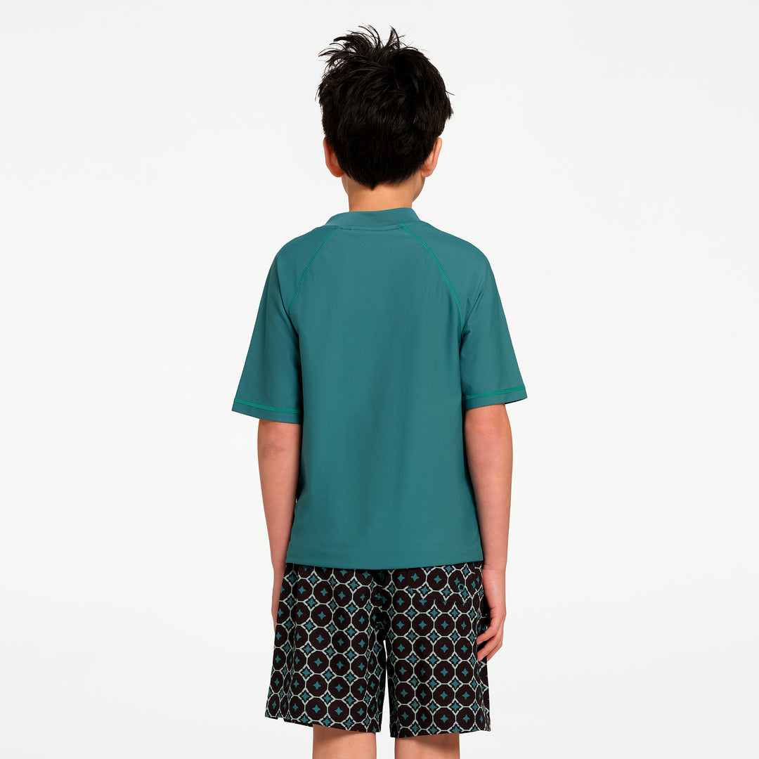 A child viewed from the back wearing a teal short-sleeve rash guard and patterned swim shorts against a plain white background.