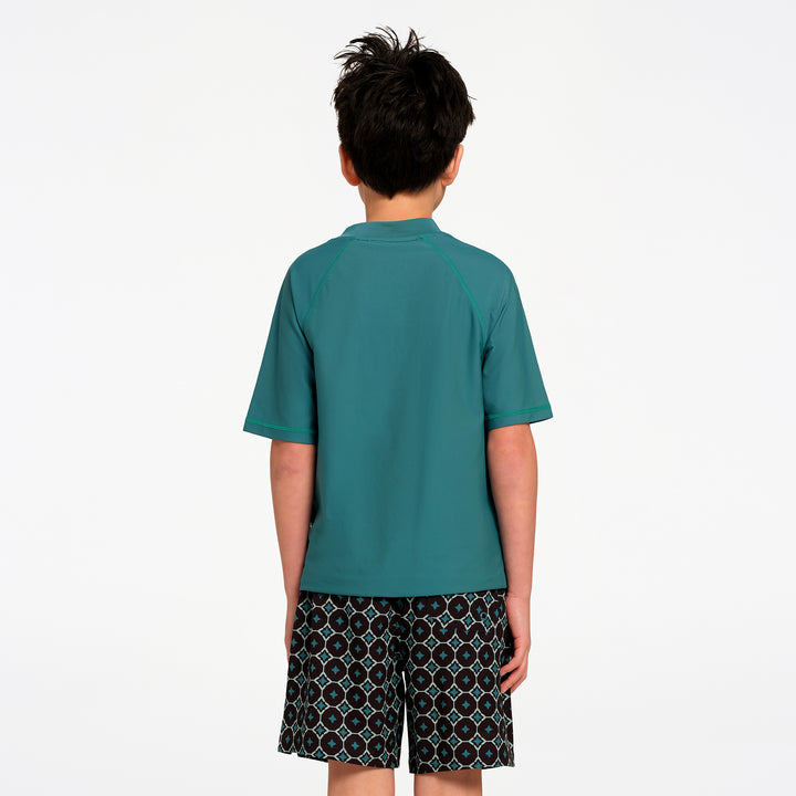 A child viewed from the back wearing a teal short-sleeve rash guard and patterned swim shorts against a plain white background.