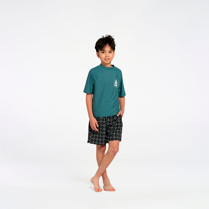 A young boy stands barefoot against a seamless white studio background, wearing a teal short-sleeve rash guard with a small white chest logo and black patterned swim shorts.