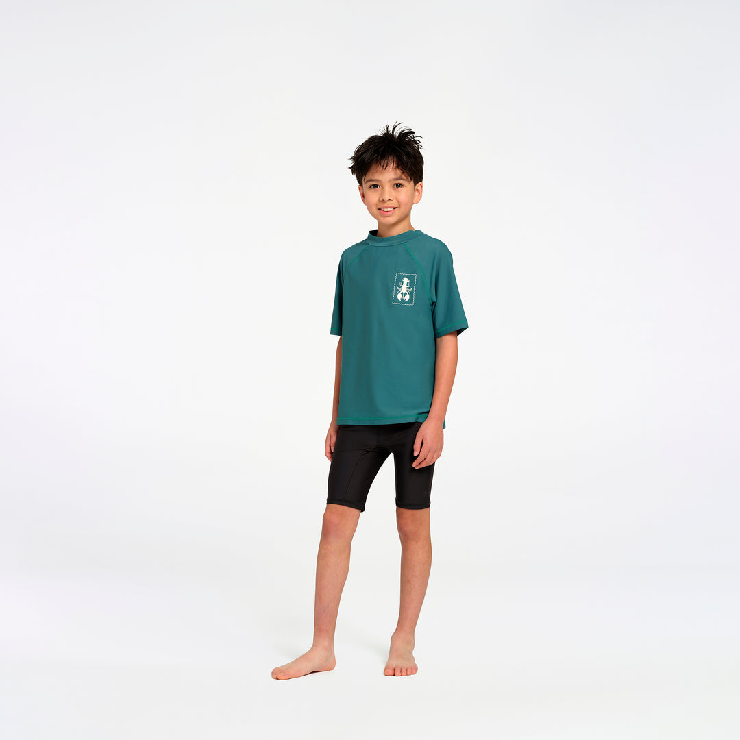 A young boy stands barefoot against a seamless white studio background, wearing a teal short-sleeve rash guard with a small white graphic on the left chest and black swim shorts.