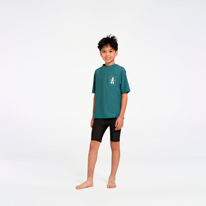 A young boy stands barefoot against a seamless white studio background, wearing a teal short-sleeve rash guard with a small white graphic on the left chest and black swim shorts.