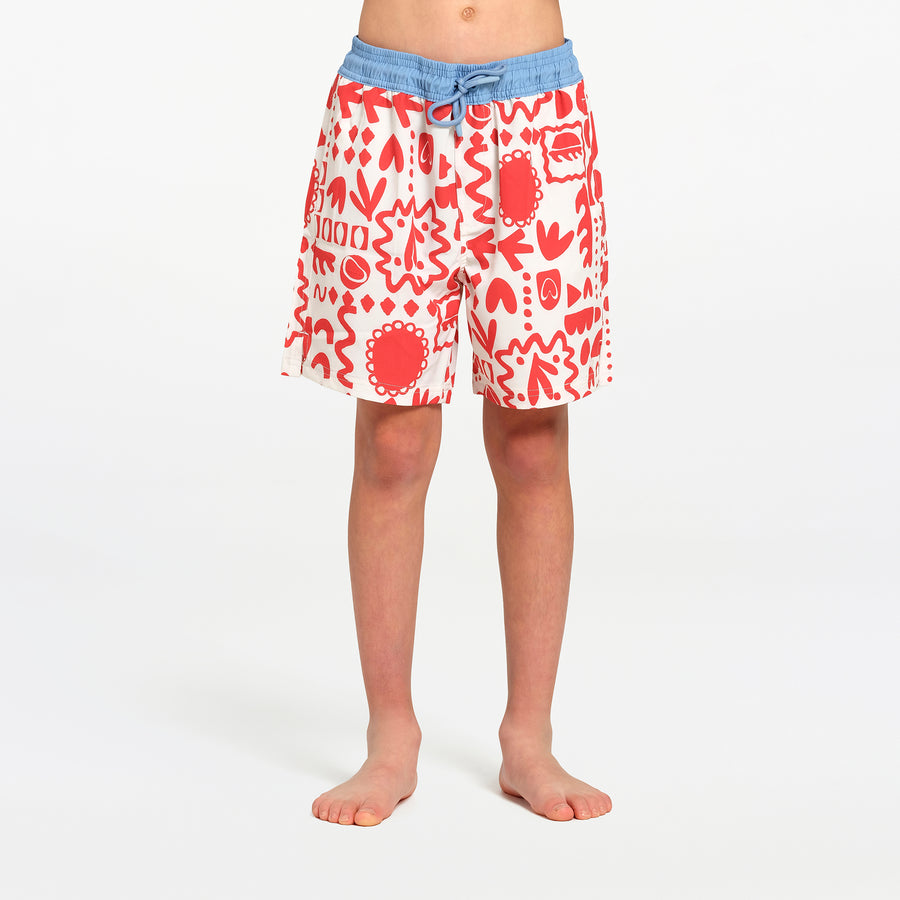 A child wearing patterned swim board shorts with a blue waistband, standing barefoot against a plain white studio background.