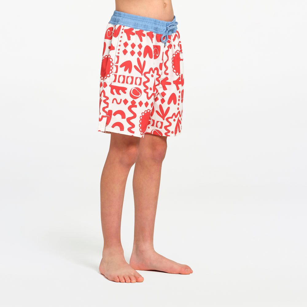 A child wearing white swim shorts with a red abstract pattern and a light blue  waistband, standing barefoot against a plain white studio background.