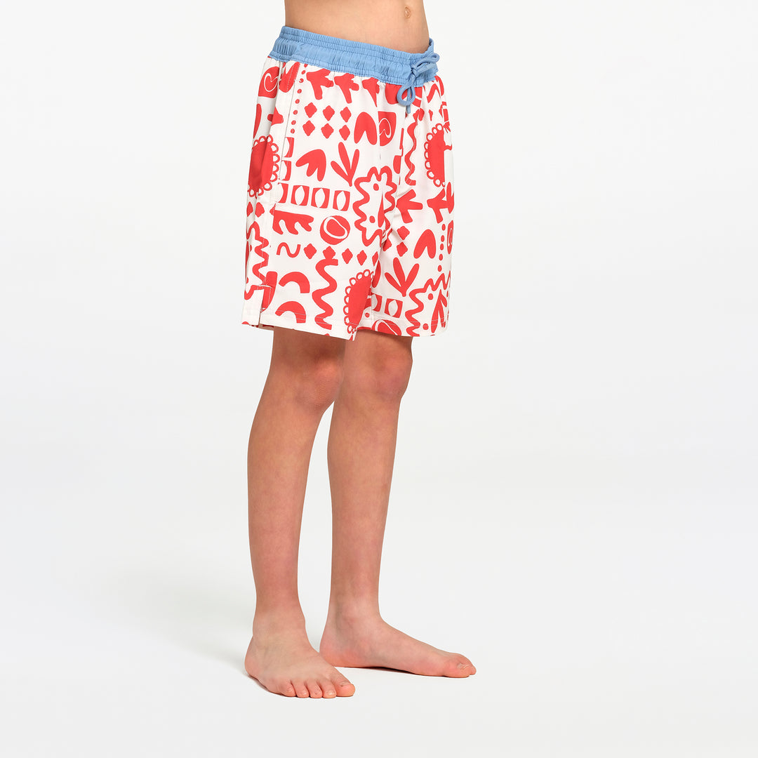 A child wearing white swim shorts with a red abstract pattern and a light blue  waistband, standing barefoot against a plain white studio background.