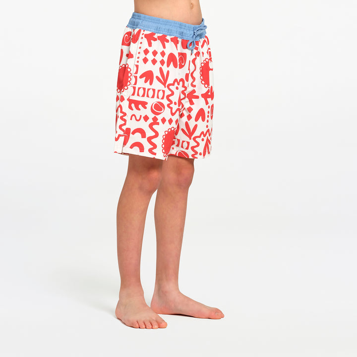 A child wearing white swim shorts with a red abstract pattern and a light blue  waistband, standing barefoot against a plain white studio background.