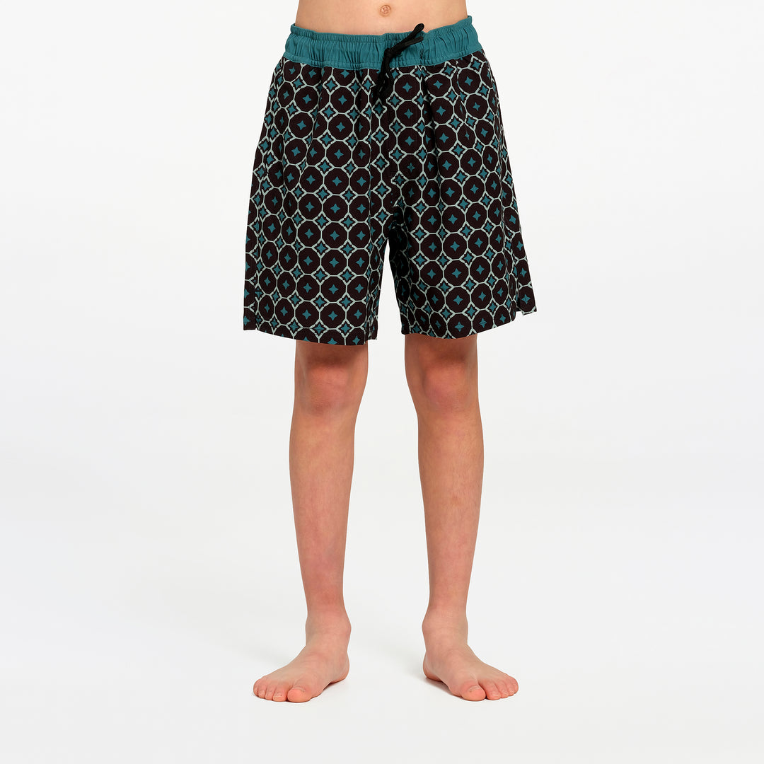 A child wearing patterned swim boardshorts with a teal waistband, standing barefoot against a plain white studio background.