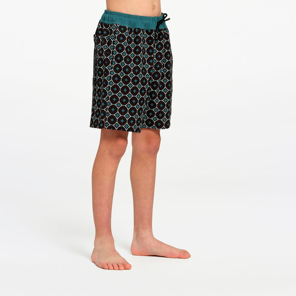 A child wearing patterned swim boardshorts with a teal waistband, standing barefoot against a plain white studio background.