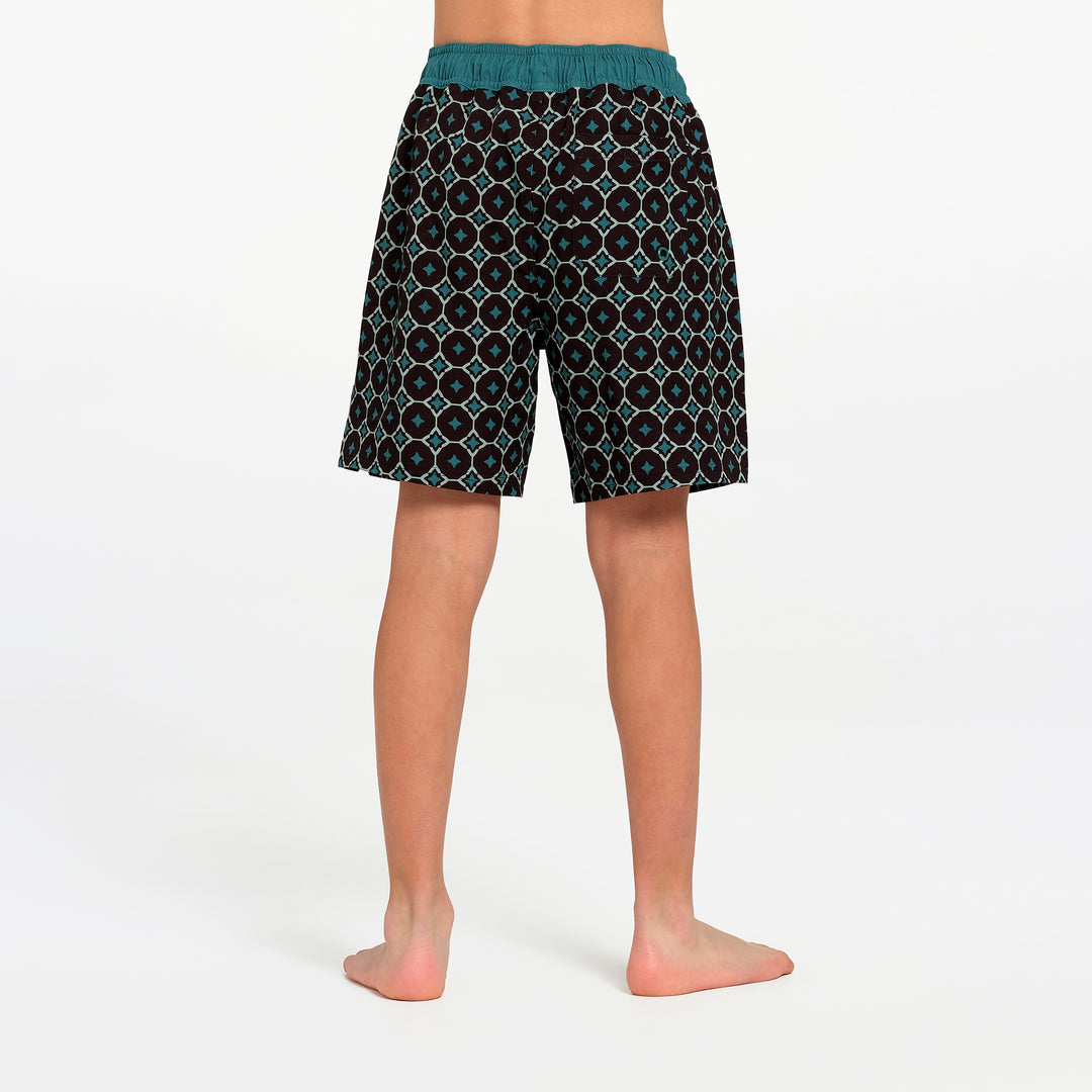 A person shown from the back wearing patterned swim board shorts with a teal waistband, standing barefoot against a clean white studio background.