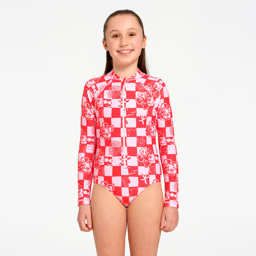 A young girl stands against a plain light gray studio background wearing a long-sleeve one-piece swimsuit with a red and white checkerboard pattern and white floral graphics.