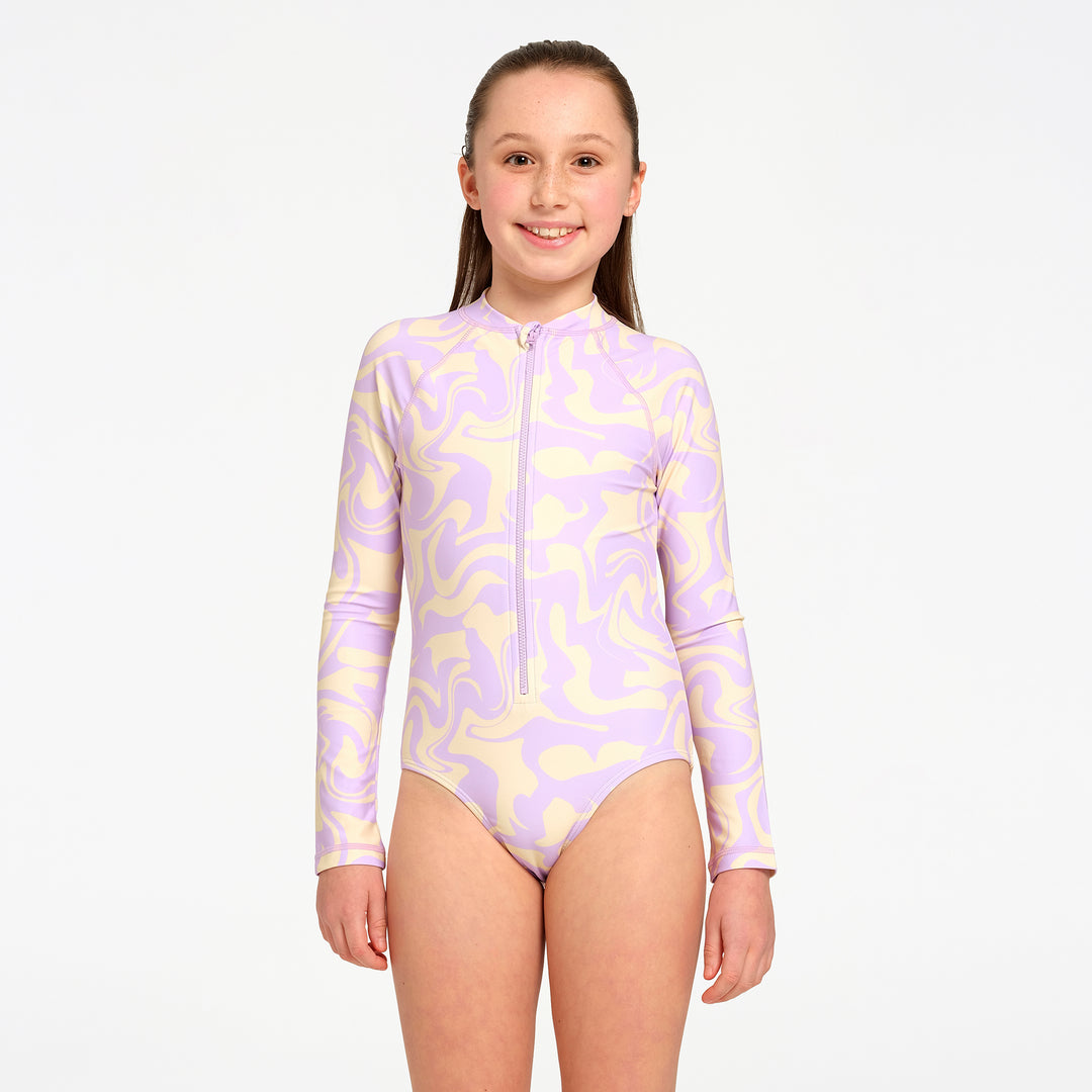 A young girl modeling a long-sleeve one-piece swimsuit with a lavender and pale yellow abstract swirl pattern and a front zipper, set against a plain white studio background.