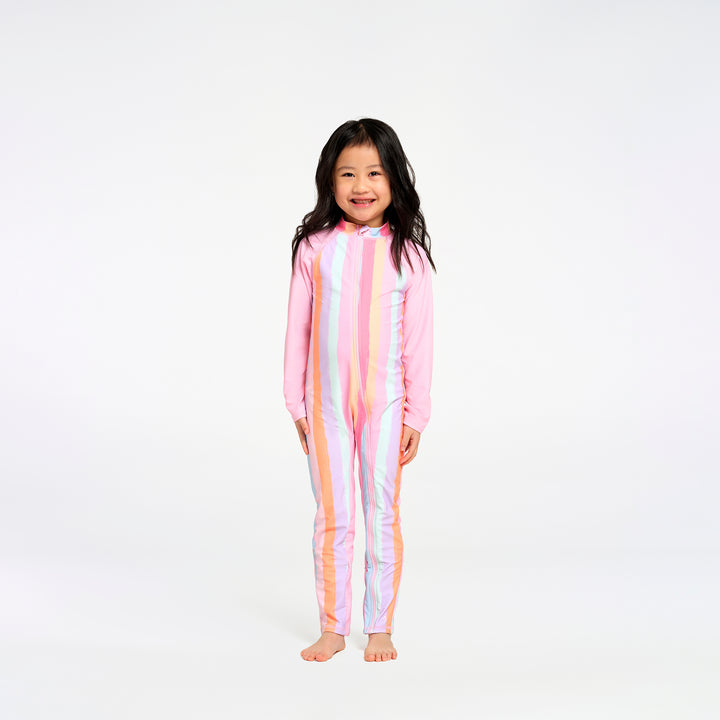 A young girl with long dark hair stands barefoot against a plain white studio background, wearing a one-piece long-sleeve swimsuit with vertical pastel stripes
